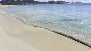 A beach with clear water, a sailboat, and mountains in the background.