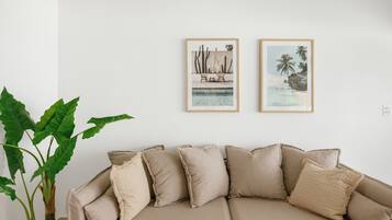 A beige sofa with multiple cushions, a large green plant, and two framed pictures on the wall.