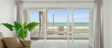 A room with a large sliding glass door leading to a beach view, a beige sofa, a large green plant, and a wicker lamp.