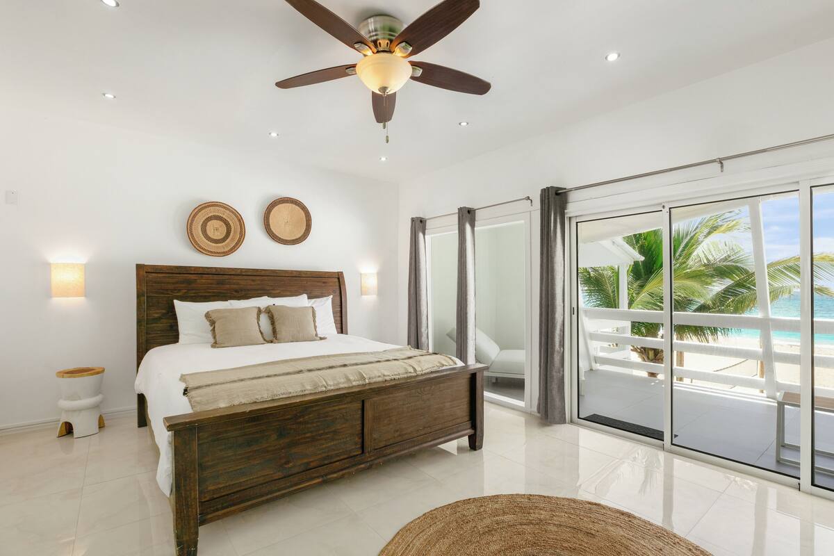 A bedroom with a wooden bed, a ceiling fan, a large window with a view of the beach, and a white toilet.