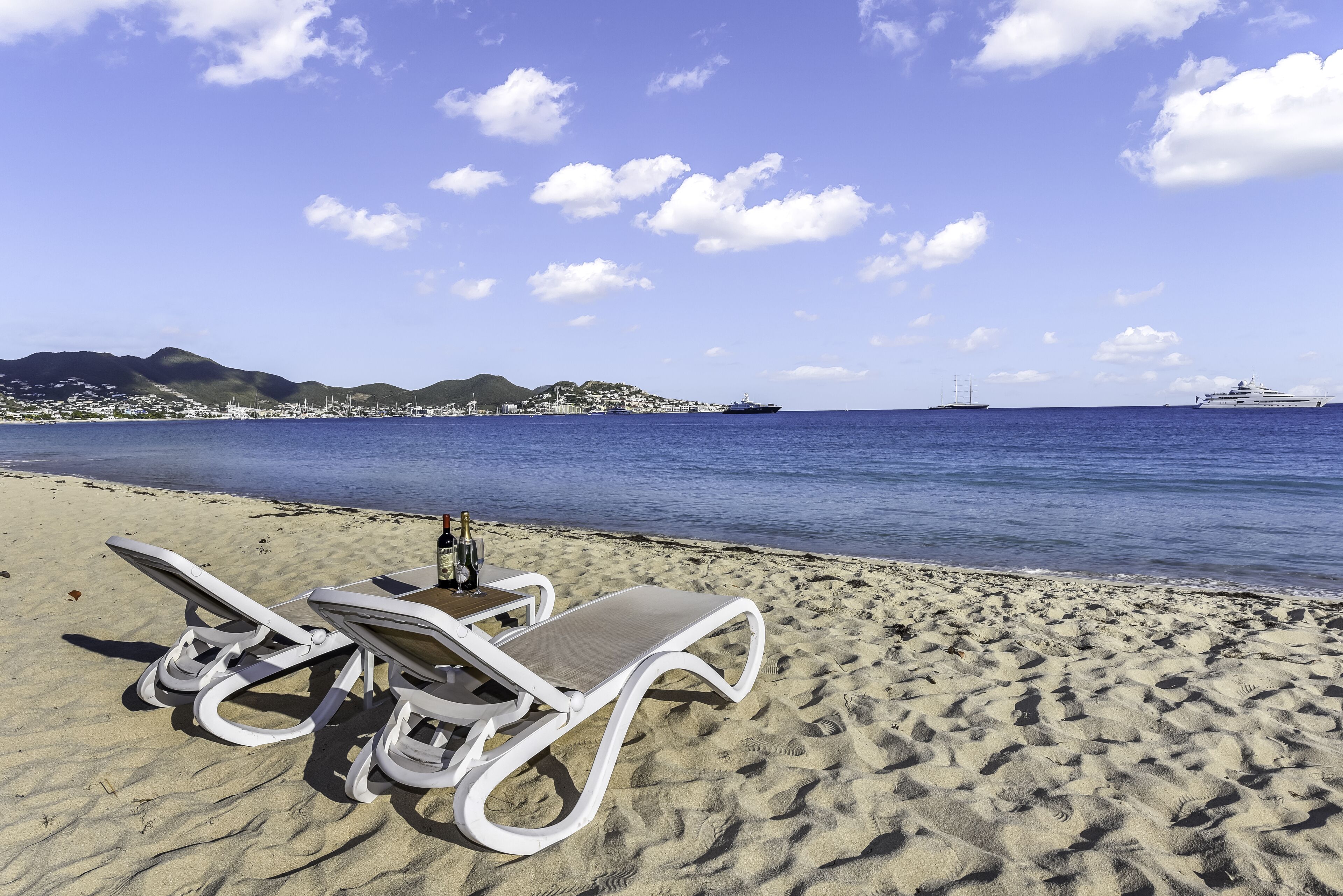 Una playa con dos sillas de playa blancas y una mesa con botellas y vasos, con vista a un mar tranquilo y un horizonte urbano distante.