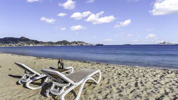 Una playa con dos sillas de playa blancas y una mesa con botellas y vasos, con vista a un mar tranquilo y un horizonte urbano distante.