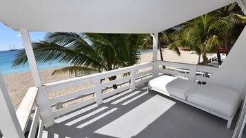 Un balcón techado con barandilla blanca, un sofá blanco y vistas a una playa de arena con palmeras y el océano.