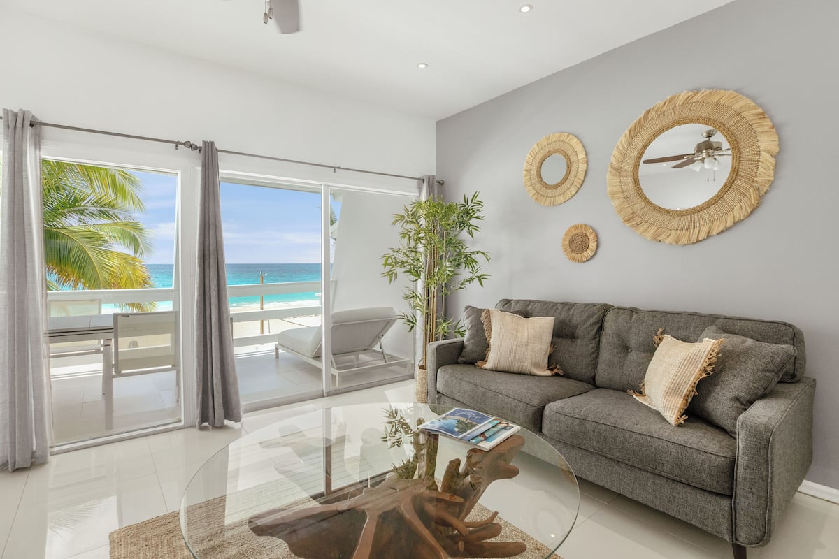 A modern living room with a grey sofa, a glass coffee table, and a view of the beach through large sliding doors.