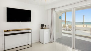 A modern hotel room with a large TV, a small fridge, a console table, and a sliding glass door leading to a beach view. - Cocovaa (Simpson Bay)