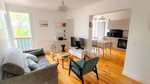 A modern living room with a sofa, coffee table, and TV. A kitchen is visible in the background with a dining area. - La riviéra" apartment. (HOSSEGOR)