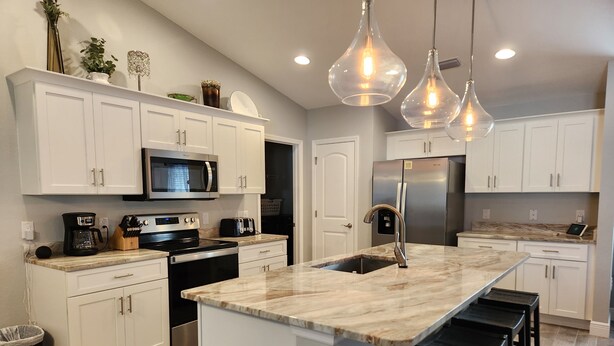 A modern kitchen with white cabinets, a central island with bar stools, stainless steel appliances, and pendant lighting. - Cape Coral Pool/Spa Home (Cape Coral)