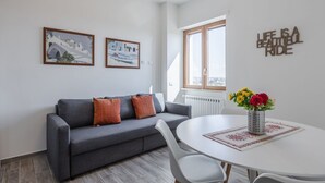 Un salon moderne avec un canapé gris, des chaises blanches, une table ronde ornée d’un vase de fleurs et deux tableaux encadrés accrochés au mur.