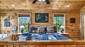 A cabin interior with a bed, two windows, a ceiling fan, a framed picture, and a sign on the wall. - Family Retreat - HotTub! Theater! FirePit! Wifi! Grill! Jacuzzi in both bedrooms (Pigeon Forge)