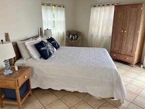 A neatly made bed with a white quilt, a wooden nightstand with a lamp, a chair, and a wooden wardrobe. - 5 bedroom Villa with pool, walk to the beach (St Thomas Parish)