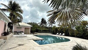 A swimming pool with a curved edge, surrounded by palm trees and a building with a pink facade. - Beautiful, centrally located, short drive to beaches and stores. (George Town)