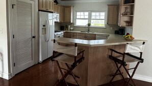 A kitchen with wooden cabinets, a refrigerator, a stove, and a sink. There are two bar stools with white cushions. - Beautiful, centrally located, short drive to beaches and stores. (George Town)