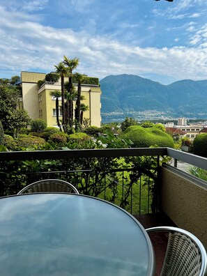 Un balcón con una mesa y sillas de vidrio, con vistas a una cordillera y un pueblo.