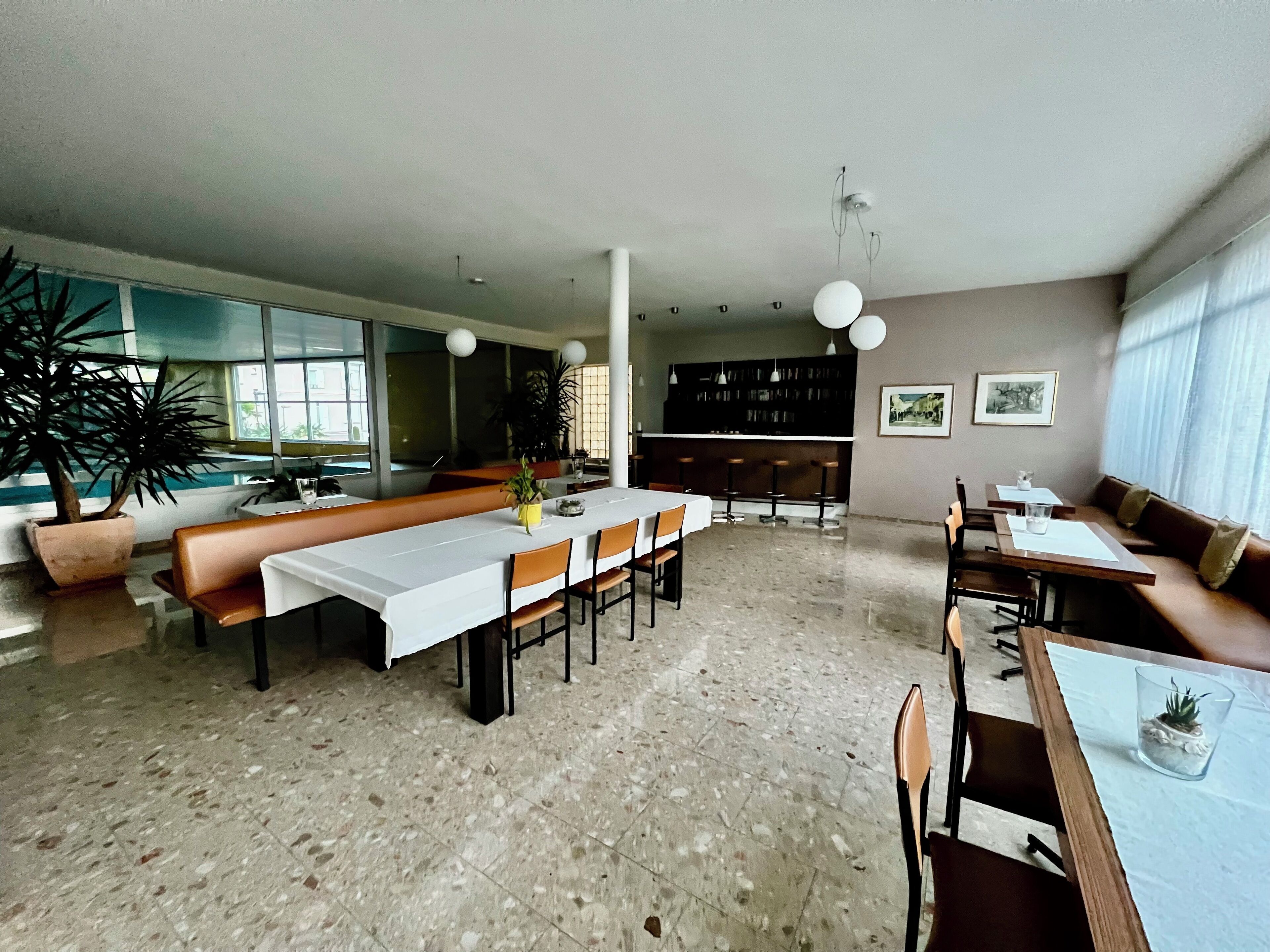 A spacious dining area with a long table, multiple chairs, and a bar counter. There are potted plants and a large window providing natural light.