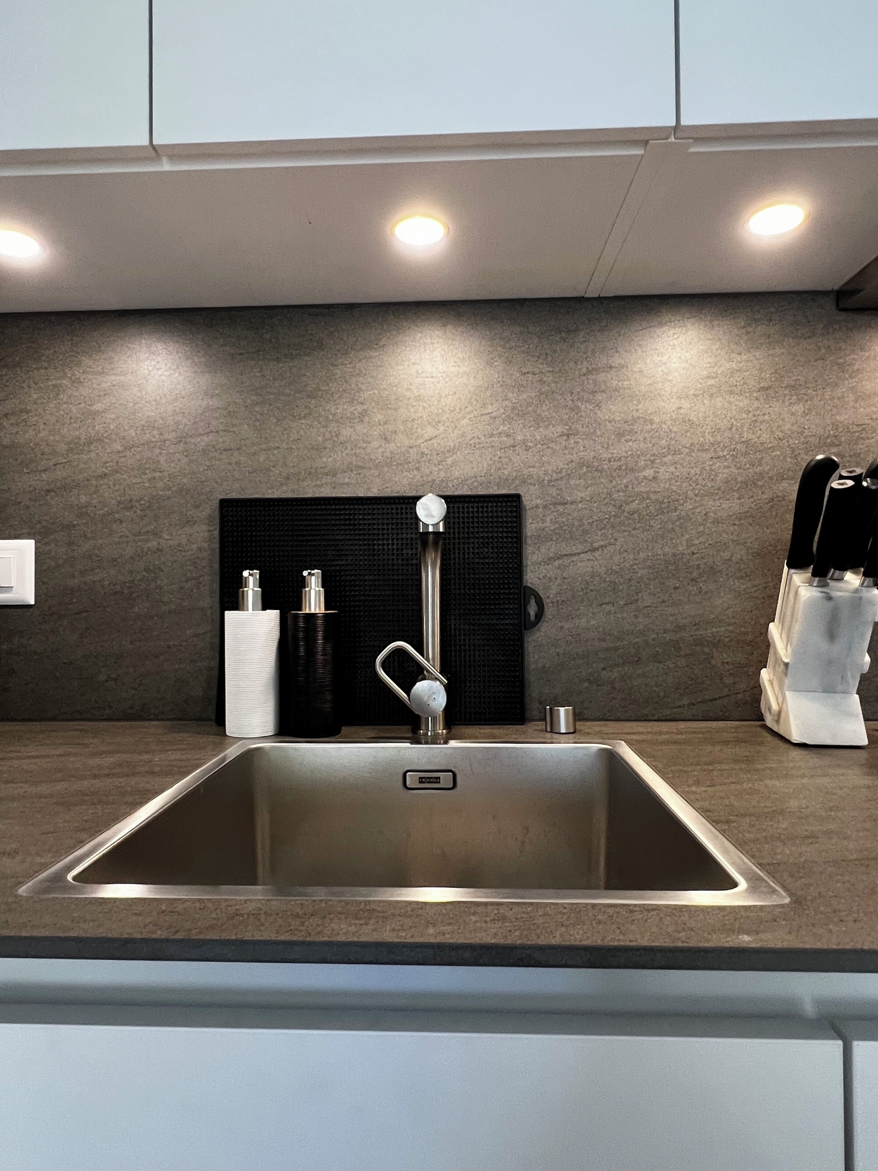 Een moderne keuken met een RVS spoelbak, een zwart gestructureerd spatwandje en inbouwspots.
