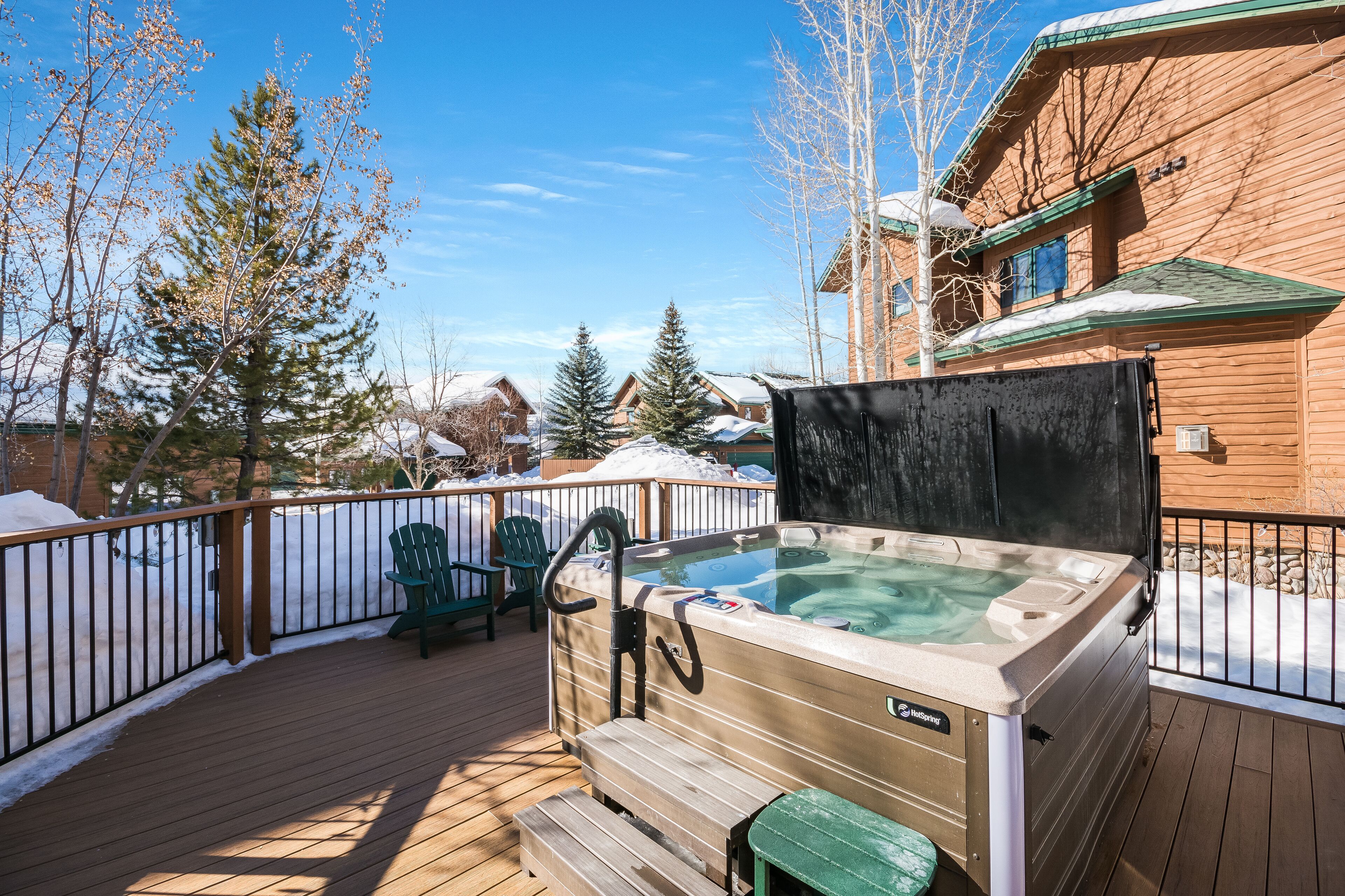 Eine Holzterrasse mit einem Whirlpool, grünen Stühlen und verschneiter Umgebung.