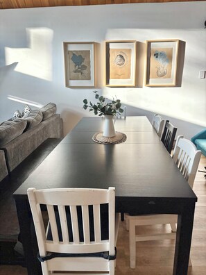 Un espace repas avec une longue table, des chaises blanches, un vase rempli de fleurs et des œuvres d’art encadrées accrochées au mur.