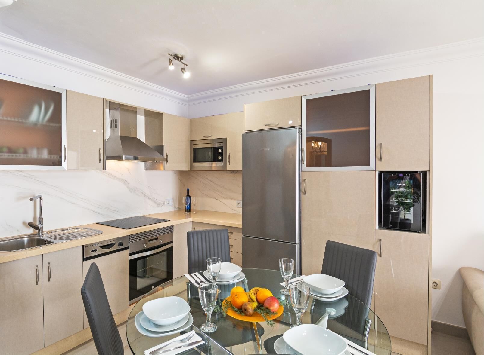 A modern kitchen with a dining area, featuring a refrigerator, oven, microwave, and a glass table set for dining.