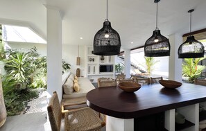 Relax in a serene space with a central dining area, wicker chairs, and a sofa, surrounded by lush greenery.