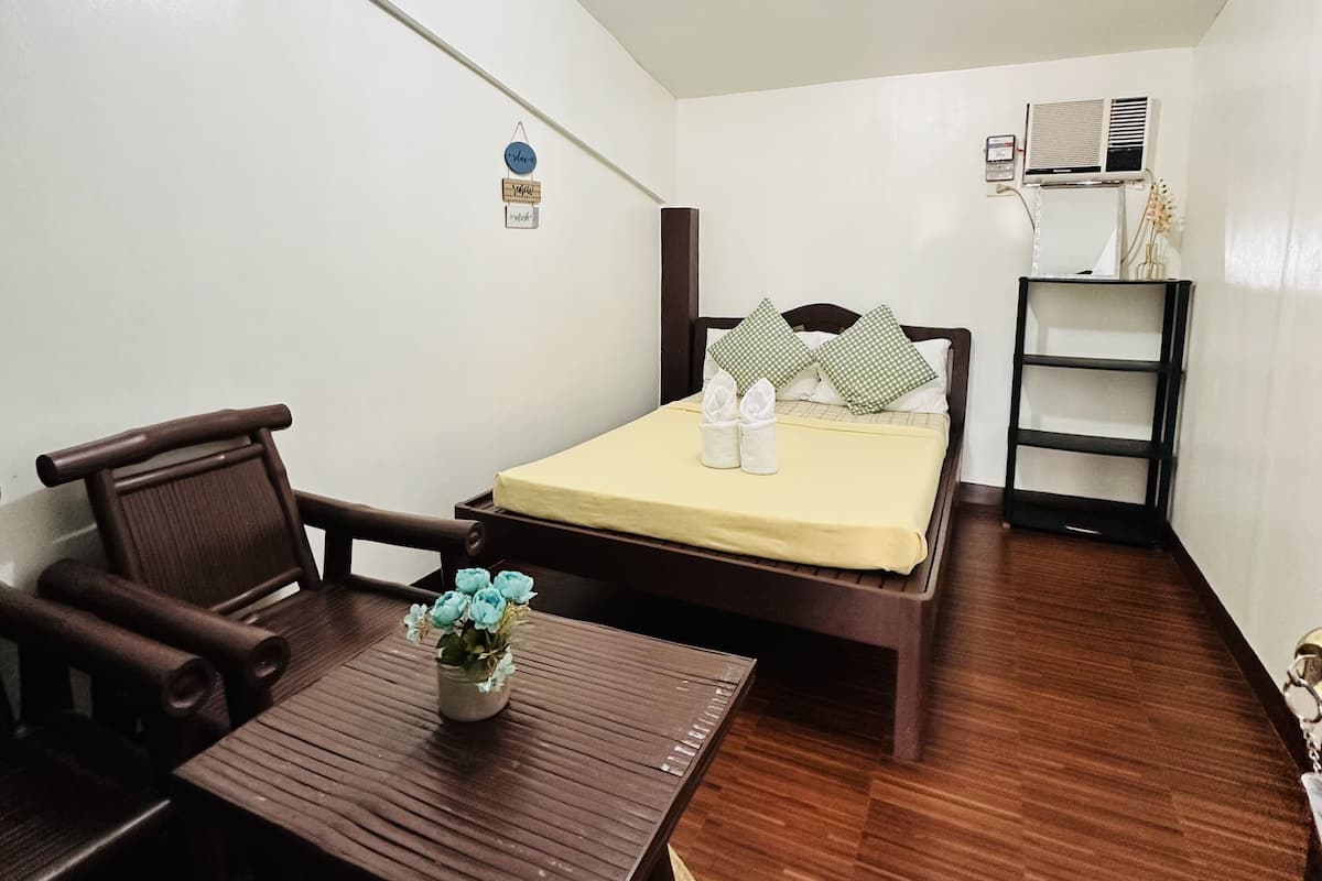 A compact room with a bed, a small table with a vase of flowers, a chair, and a shelf with an air conditioner.
