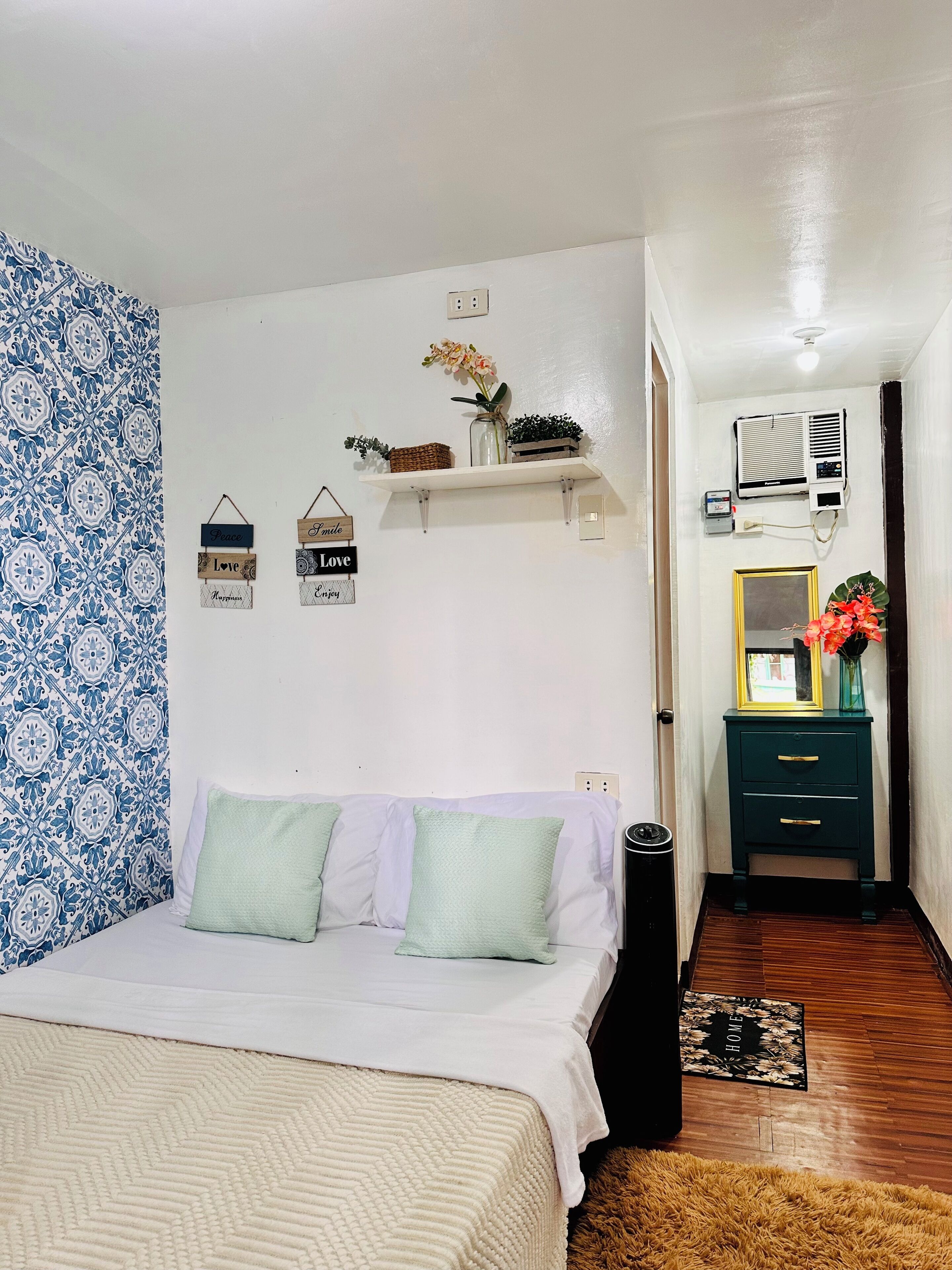 A bedroom with a bed, a nightstand, a wall-mounted shelf with decorative items, a wall outlet, and a door leading to another room.