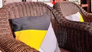 Des chaises en osier avec des coussins, un éventail rouge et un balcon doté de balustrades en bois et de plantes vertes.