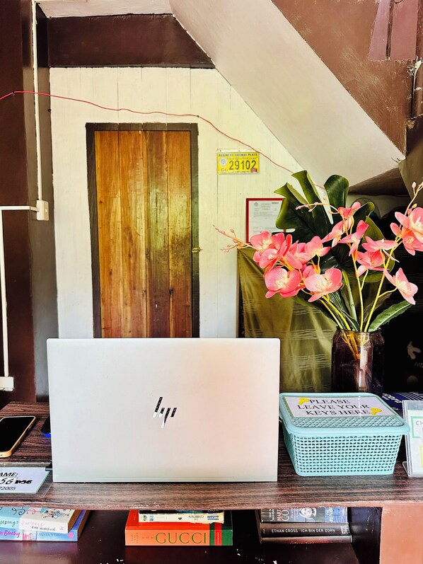 A laptop on a desk with a vase of pink flowers, a wooden door, and a sign with a number. - Amancio's Balai (Puerto Princesa)