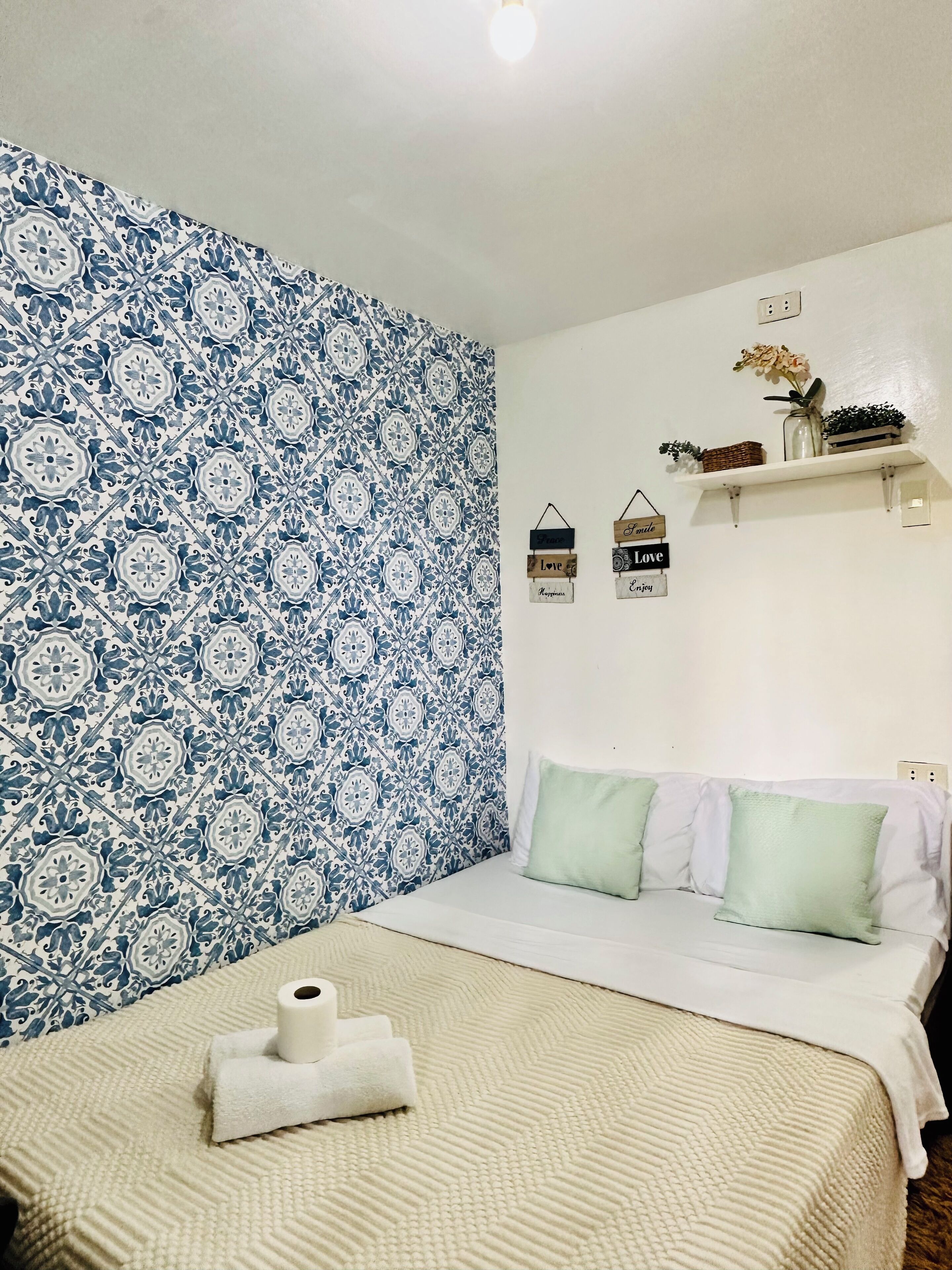 A neatly made bed with a white and green pillow set, a beige textured bedspread, and a roll of toilet paper on the bed.