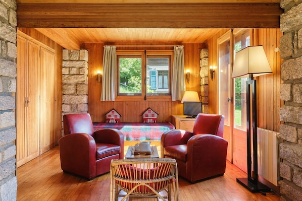 A cozy room with wooden walls, a stone fireplace, a large window with curtains, a television, and a lamp. - Petit Chalet Des Moussoux, Chamonix, France (Chamonix)