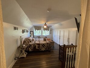 Ein schmaler Flur mit einem Bett, einem Sessel und einem Kinderbett. Weiße Wände, ein Deckenventilator und ein Fenster mit Vorhängen.