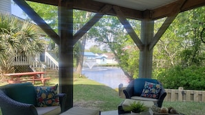 A covered patio with wicker furniture, a small table with a plant, and a view of a water body and distant buildings. - Ocean Lakes Private Beach & Fountain View Tower (Myrtle Beach)