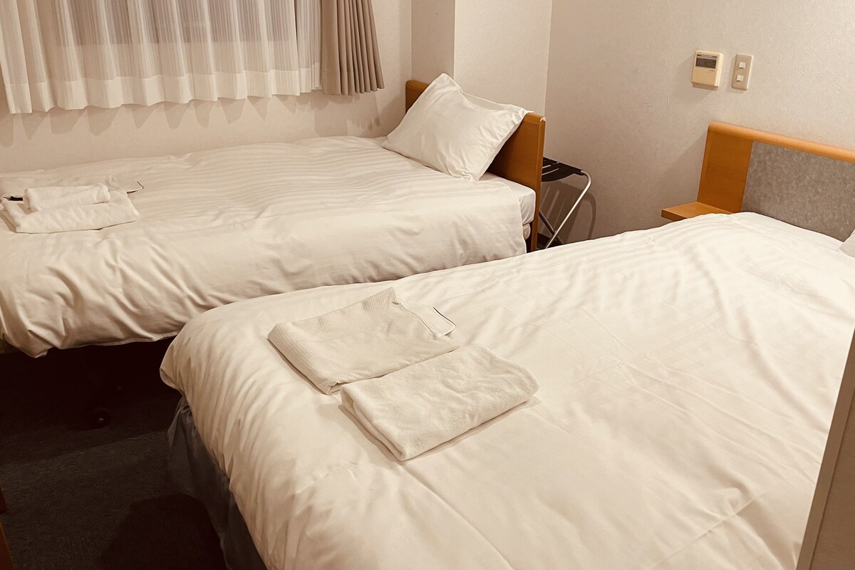 Two single beds with white linens and towels, a wooden headboard, a nightstand, and a wall-mounted thermostat.