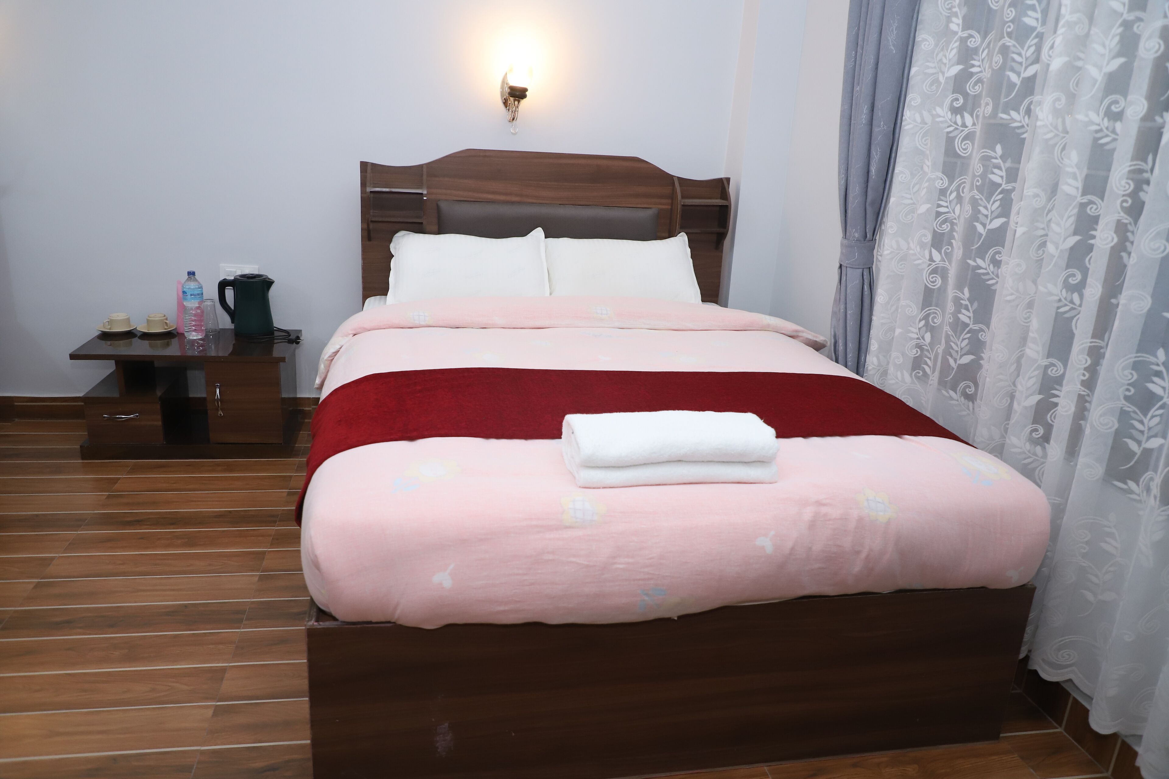 A neatly made bed with a wooden headboard, a bedside table with a lamp, and a curtain with a floral pattern.