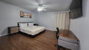 A bedroom with a bed, a desk, a chair, a nightstand, a ceiling fan, and a painting on the wall.