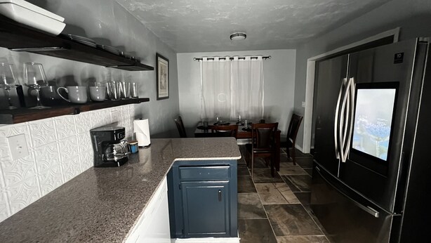 A modern kitchen with granite countertops, a coffee machine, and a built-in oven. There is a dining area with a table and chairs, and a refrigerator with a digital display.