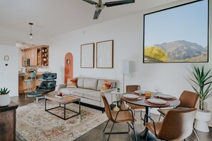 A modern living room with a sofa, coffee table, and dining area. There is a kitchen in the background with wooden cabinets and a dining table set for four.