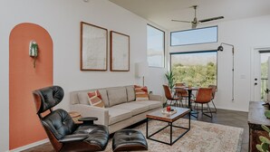 A modern living room with a sofa, leather chairs, a coffee table, and a dining area with chairs and a table. There are framed artworks on the wall and a ceiling fan.