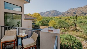 A balcony with wicker chairs and a small table, overlooking a mountainous landscape with cacti and shrubs.