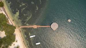 Vue aérienne d’une plage avec une jetée en bois s’étendant dans l’eau, un bateau amarré à la jetée et une plage de sable bordée de palmiers.