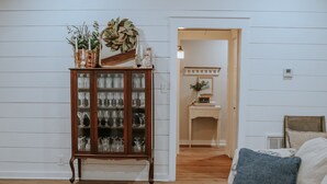 A living room with a high ceiling, wooden beams, a glass cabinet with decorative items, a beige sofa with cushions, and a white rug. - The Shanty @ Rayburn Country (Brookeland)