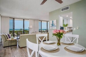 Ein modernes Wohnzimmer mit Balkon, einem Essbereich mit weißen Stühlen und Tellern, und eine durchgehende Tür sichtbare Küche.