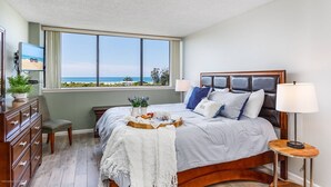 A bedroom with a large window offering a view of the beach, a bed with multiple pillows, a bedside table with a lamp, and a dresser. - Ocean Views in Our House at the Beach, Full Gulf Views from Balcony, Heated Pools! (Siesta Key)