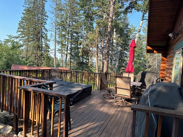 A wooden deck with outdoor furniture, including chairs and a table, surrounded by tall trees. - Deer Pass Lodge (Lead)
