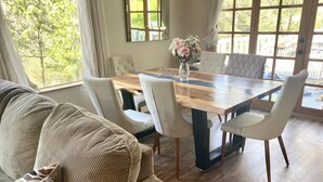 Uma sala de jantar com mesa de madeira e cadeiras estofadas brancas, um sofá bege e um vaso com flores.
