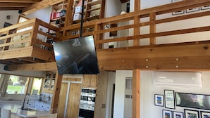 Um amplo quarto estilo loft com teto alto, vigas de madeira, uma grande TV de tela plana e uma varanda com vista para a região.