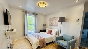 A modern bedroom with a bed, a chair, a TV, and a window. - Cozy 1-Bedroom Apartment in the Heart of Grimsby (Grimsby)