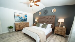 A bedroom with a tufted headboard, a large bed, two bedside tables with lamps, a ceiling fan, a wall-mounted abstract painting, and a wooden cabinet.