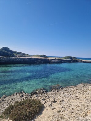 A coastal landscape with clear turquoise waters, rocky shores, and distant hills.