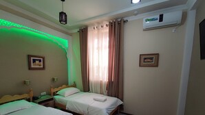 A hotel room with two beds, a green-lit wall, an air conditioner, and a window with curtains. - Naqshband Craft Center & Hotel (Bukhara)