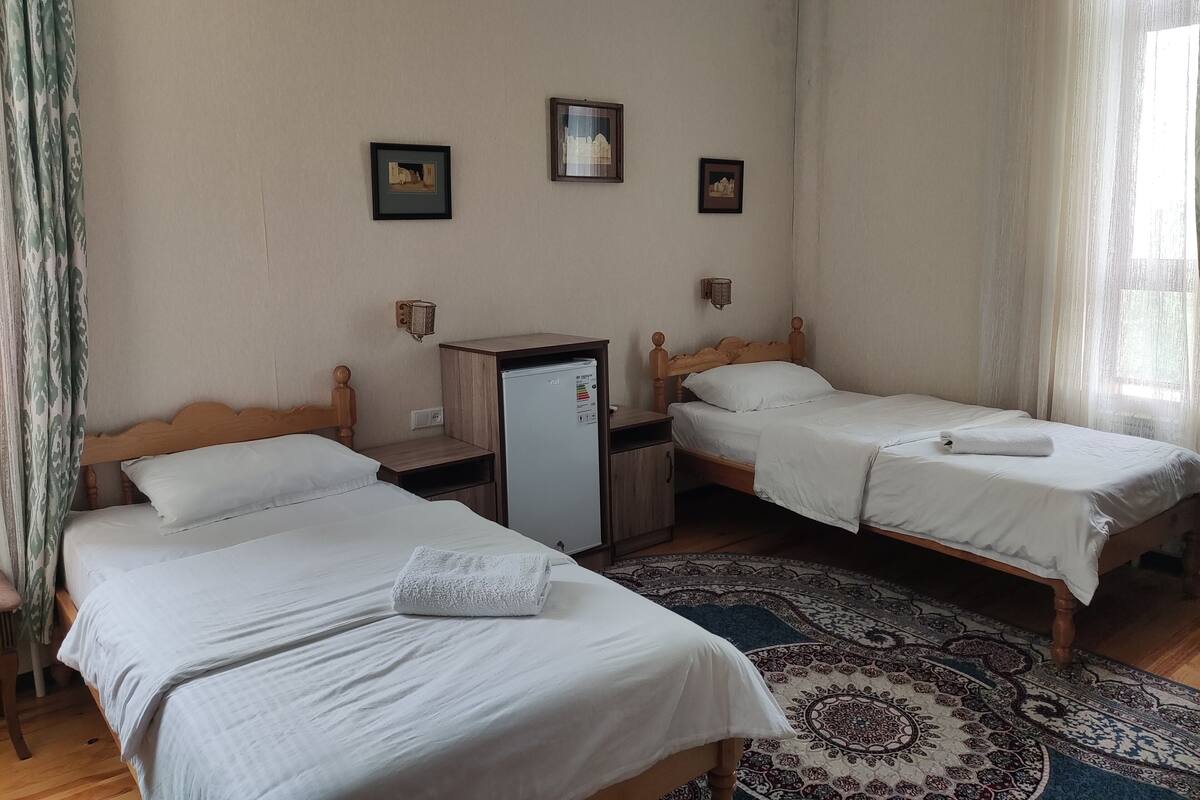 Une chambre d’hôtel avec deux lits simples, un petit bureau avec un réfrigérateur, un tapis à motifs et une fenêtre avec des rideaux légers.